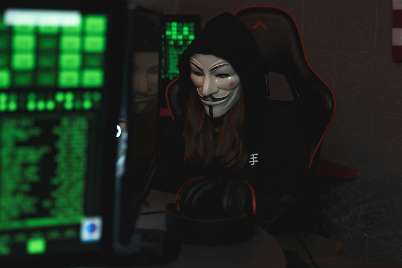 A mysterious hacker wearing a Guy Fawkes mask and black hoodie in a dimly lit room focused on computer screens.
